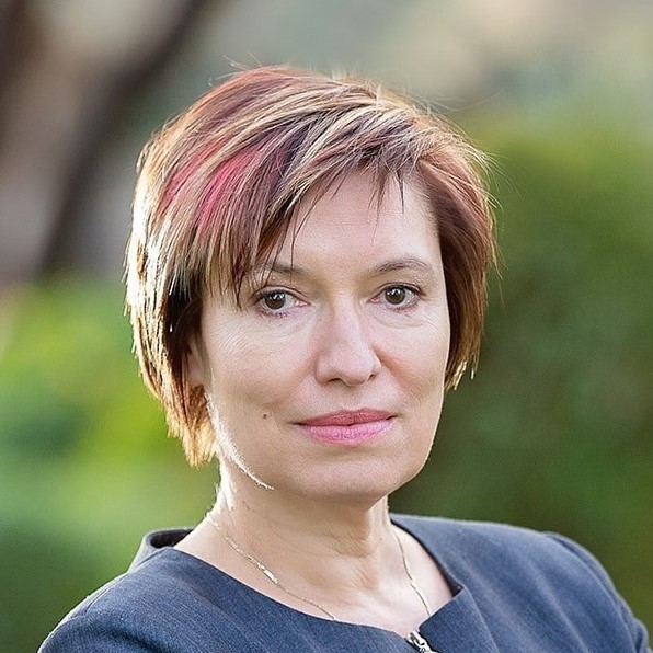 Headshot of Petya Maneva. She has short red, pink and blond hair with brown eyes. She is wearing a dark blue suit jacket. Background is blurred but has bushes and trees.